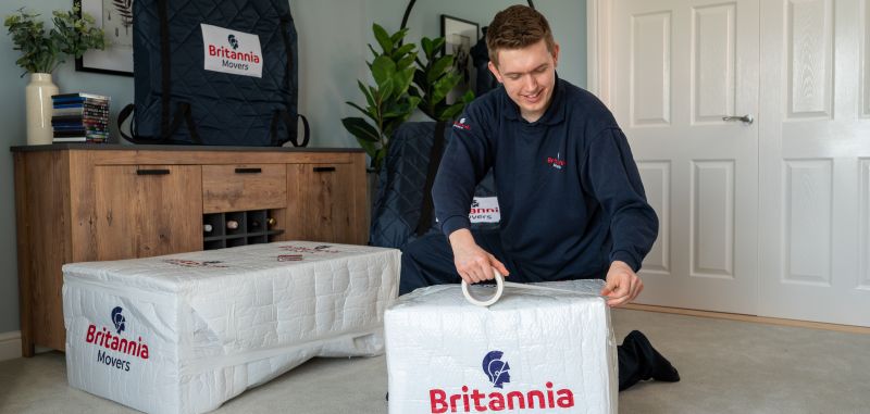 A smiling uniformed Britannia removals operative works in a lounge wrapping a side table in branded packaging ready for a international removal