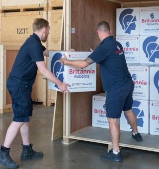 Two uniformed removal men load Britannia Robbins branded boxes into a wooden storage container inside a large, clean warehouse