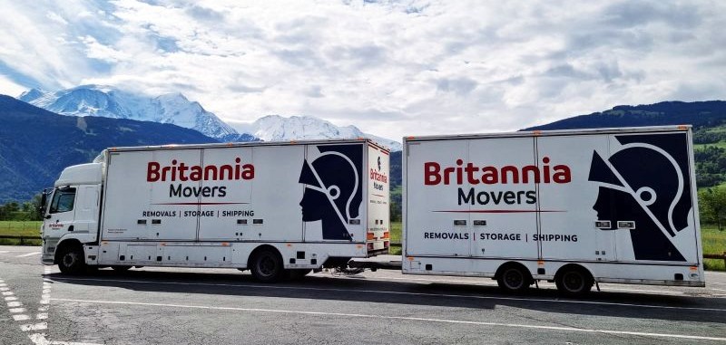 A Britannia-branded road train truck carrying out a removal in Europe, pictured on a sunny day on a road with high snow-covered mountains in the background.