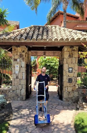 A removal man wearing Britannia uniform walks out of a tiled Spanish gateway pushing a sack truck, surrounded by palm trees and flowers. 