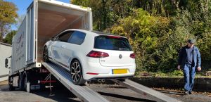 A white car is loaded into a Britannia-branded shipping container on ramps, overseen by a uniformed staff member