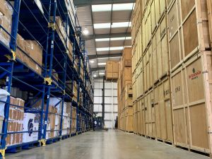 A forklift truck lifts a wooden storage crate onto a stack in a large, clean warehouse, with hundreds of Britannia branded crates and boxes on both sides. 